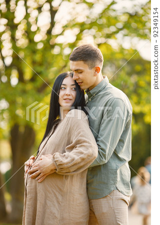 Pregnant woman and her husband standing in a park on a grass and hugging 92349154