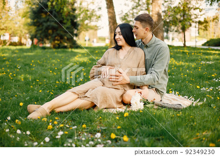 Pregnant woman and her husband sitting in a park on a grass and hugging 92349230