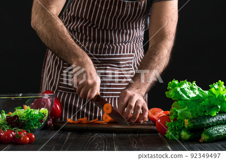 Young home cook man in apron slicing carrot with kitchen knife. 92349497