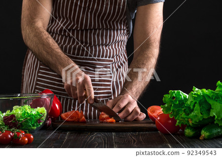Man preparing salad on a wooden table. Men's hands cut the tomato to make a salad on black background Man preparing salad on a wooden table. Men's hands cut the tomato to make a salad on black background 92349543