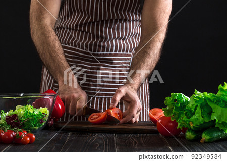 Man preparing salad on a wooden table. Men's hands cut the tomato to make a salad on black background 92349584