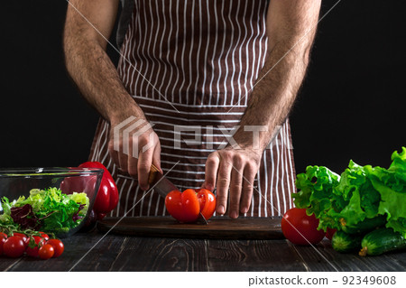Man preparing salad on a wooden table. Men's hands cut the tomato to make a salad on black background Man preparing salad on a wooden table. Men's hands cut the tomato to make a salad on black background 92349608
