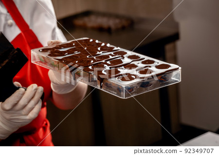A woman confectioner with red uniform and white sterile gloves do a set of colorful chocolates from milk chocolate on a table. A woman confectioner with red uniform and white sterile gloves do a set of colorful chocolates from milk chocolate on a table. 92349707