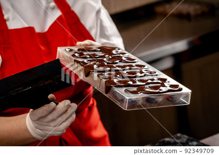 A woman confectioner with red uniform and white sterile gloves do a set of colorful chocolates from milk chocolate on a table. A woman confectioner with red uniform and white sterile gloves do a set of colorful chocolates from milk chocolate on a table. 92349709