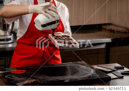 A woman confectioner with red uniform and white sterile gloves do a set of colorful chocolates from milk chocolate on a table. A woman confectioner with red uniform and white sterile gloves do a set of colorful chocolates from milk chocolate on a table. 92349711