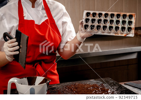 A woman confectioner with red uniform and white sterile gloves do a set of colorful chocolates from milk chocolate on a table. 92349715