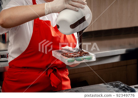 A woman confectioner with red uniform and white sterile gloves do a set of colorful chocolates from milk chocolate on a table. 92349776