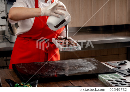A woman confectioner with red uniform and white sterile gloves do a set of colorful chocolates from milk chocolate on a table. A woman confectioner with red uniform and white sterile gloves do a set of colorful chocolates from milk chocolate on a table. 92349782