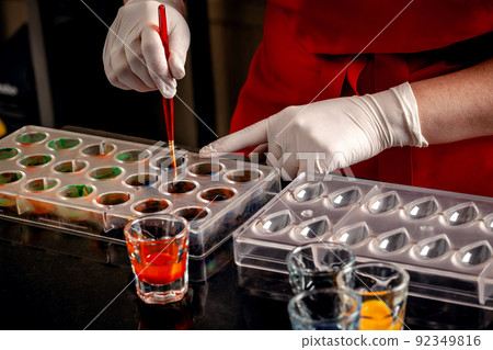 A woman confectioner with red uniform and white sterile gloves do a set of colorful chocolates from milk chocolate on a table. 92349816
