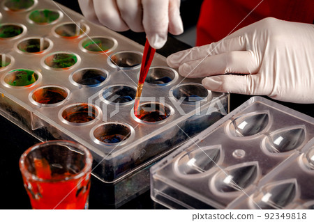 A woman confectioner with red uniform and white sterile gloves do a set of colorful chocolates from milk chocolate on a table. A woman confectioner with red uniform and white sterile gloves do a set of colorful chocolates from milk chocolate on a table. 92349818