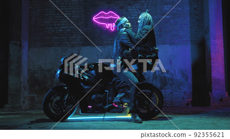 A girl in love and a guy are sitting on a super sport motorcycle flirting and hugging against the background of a neon sign A girl in love and a guy are sitting on a super sport motorcycle flirting and hugging against the background of a neon sign 92355621