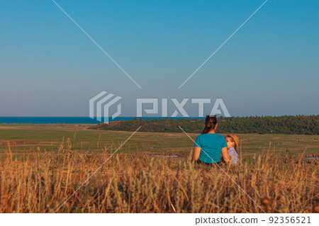 Mother and daughter sitting by lakes nature lanscape. A little girl with long blond hair braid hugging with mom outdoor. Mother and daughter sitting by lakes nature lanscape. A little girl with long blond hair braid hugging with mom outdoor. 92356521
