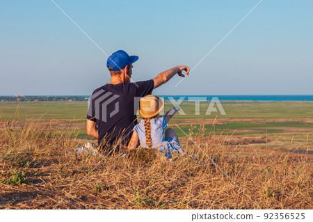Father and daughter sitting by lakes on a nature lanscape. A little girl with long blond hair braid hugging dad outdoor. 92356525