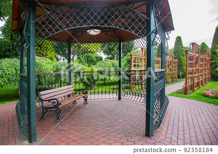 forged iron gazebo with ceiling lamp with white diffuser, place for relaxing on wooden bench in backyard with vertical pergola for growing climbing flowers in garden with bushes and trees, nobody. forged iron gazebo with ceiling lamp with white diffuser, place for relaxing on wooden bench in backyard with vertical pergola for growing climbing flowers in garden with bushes and trees, nobody. 92358184