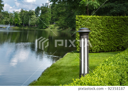 garden iron lantern of backyard lighting in trimmed boxwood bushes near lawn meadow on background pond with water and coast with trees, green landscaping of scenic, eco friendly theme, nobody. garden iron lantern of backyard lighting in trimmed boxwood bushes near lawn meadow on background pond with water and coast with trees, green landscaping of scenic, eco friendly theme, nobody. 92358187