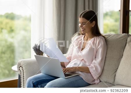 Attractive woman sorting out papers sit on sofa use laptop Attractive woman sorting out papers sit on sofa use laptop 92362209