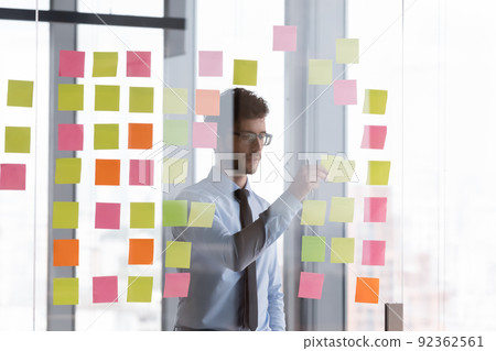 Focused millennial businessman sticking paper notes on glass wall 92362561