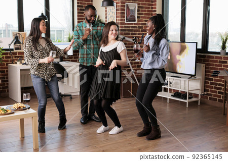Multicultural group of close friends dancing together at wine party. Joyful young people having fun in living room while celebrating friendship anniversary event with music and alcoholic beverages. 92365145