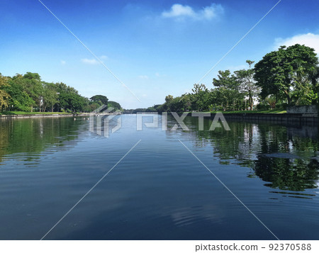 A large pool in the middle of the park is filled with large trees surrounded by the bright blue sky 92370588