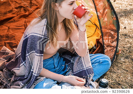 Happy young girl sitting in tent drinking hot tea from cup of thermos against backdrop of spring or fall forest. Mental health. Time for yourself, contemplation, meditation. Happy young girl sitting in tent drinking hot tea from cup of thermos against backdrop of spring or fall forest. Mental health. Time for yourself, contemplation, meditation. 92398626
