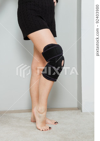 Black knee support brace on a woman leg. girl in an orthosis in the interior of the house. Orthopedic anatomic braces for knee fixation, injuries and pain. Knee Joint Bandage Sleeve. Elastic Sports. Black knee support brace on a woman leg. girl in an orthosis in the interior of the house. Orthopedic anatomic braces for knee fixation, injuries and pain. Knee Joint Bandage Sleeve. Elastic Sports. 92403980