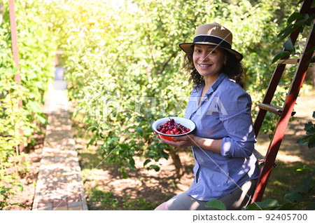 Beautiful woman, eco farmer smiling toothy smile, looking at camera, holding a bowl with freshly picked ripe cherries 92405700
