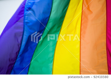 Close Up rainbow gay pride flag outside on a street. Symbol of the Lesbian Bisexual Transgender LGBT community waving in wind against cloudy sky. Social movement for freedom and equliaty. Copy Space 92410380