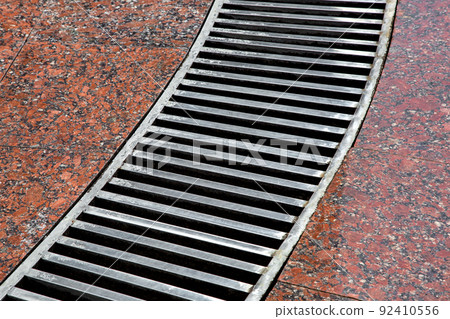 curved drainage system with stripes grate on the granite floor with a wet red stone granite surface, close-up of a storm system wet after rain weather, nobody. 92410556