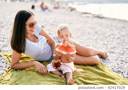 Smiling mom holding out peach to little girl eating watermelon on beach Smiling mom holding out peach to little girl eating watermelon on beach 92417029