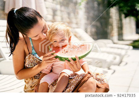Mom feeds a small baby in her arms with a piece of ripe watermelon Mom feeds a small baby in her arms with a piece of ripe watermelon 92417035