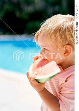 Little girl eats a watermelon, holding it with her hands. Side view 92417158
