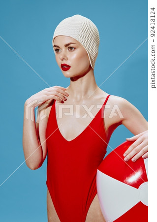A swimmer in red looks into the distance. Conceptual retro photo of a beautiful young woman in a swimsuit with a ball on a blue background 92418124