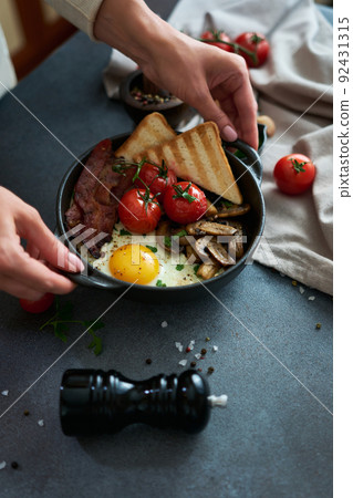Fried eggs, bacon, tomato and toasted bread in a black ceramic pan at domestic kitchen 92431315