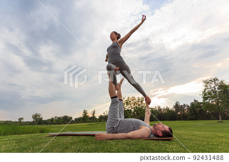 Healthy man lying on grass and balancing woman. Couple doing acrobatic yoga exercise in park Healthy man lying on grass and balancing woman. Couple doing acrobatic yoga exercise in park 92431488