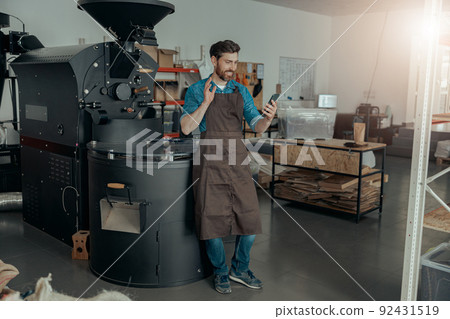 Male barista making video call standing on the background of coffee roast machine 92431519