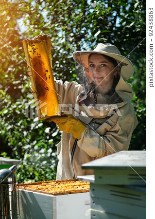 Young female beekeeper hold wooden frame with honeycomb. Collect honey. Beekeeper on apiary. Beekeeping concept. Young female beekeeper hold wooden frame with honeycomb. Collect honey. Beekeeper on apiary. Beekeeping concept. 92438863