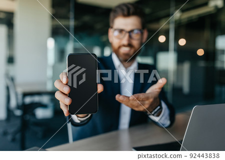 Happy businessman showing smartphone with blank screen, sitting at modern office, mockup for mobile app or website 92443838