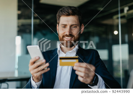 Online banking concept. Happy caucasian general manager making online payment on smartphone, sitting in office 92443990