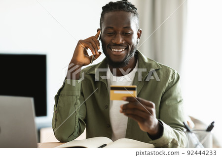 Excited black man holding credit card and calling his bank or webstore manager, working on laptop at home 92444233