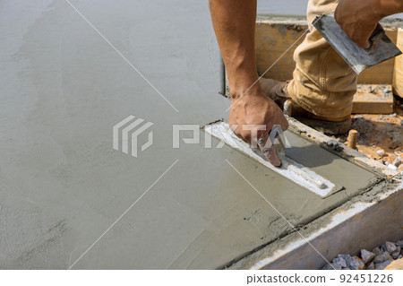 A masonry worker is smoothing the plastering concrete to a cement floor while holding a steel trowel in his hand A masonry worker is smoothing the plastering concrete to a cement floor while holding a steel trowel in his hand 92451226