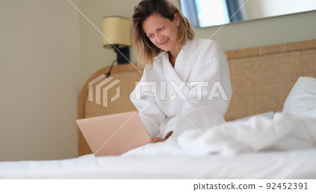 Woman sits at home on bed in white bathrobe and works on laptop Woman sits at home on bed in white bathrobe and works on laptop 92452391