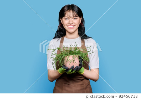 Smiling young woman in apron gloves with potted plant on blue isolated background Smiling young woman in apron gloves with potted plant on blue isolated background 92456718