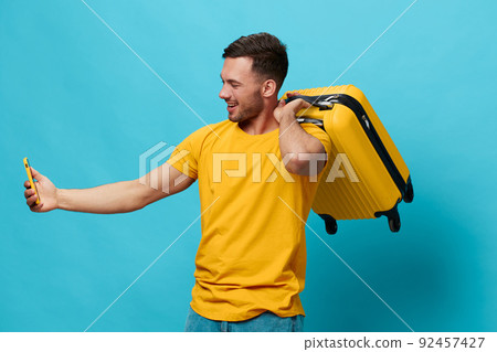 Happy cheerful tanned handsome man in yellow t-shirt doing selfie video call with phone hold suitcase posing isolated on blue studio background. Copy space Banner Mockup. Trip journeys concept 92457427