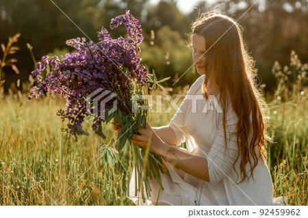 Young beautiful woman, wearing white dress, holding flowers on the meadow. Girl joying nature and freedom. Natural beauty. Stress free, dreaming. Sunset. 92459962