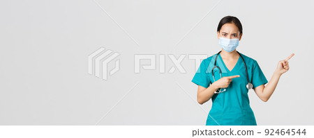 Covid-19, coronavirus disease, healthcare workers concept. Frowning suspicious or frustrated asian female doctor, wear medical mask and scrubs, pointing upper right corner, white background Covid-19, coronavirus disease, healthcare workers concept. Frowning suspicious or frustrated asian female doctor, wear medical mask and scrubs, pointing upper right corner, white background 92464544