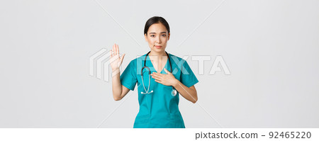 Covid-19, healthcare workers and preventing virus concept. Smiling honest asian female doctor, intern in scrubs giving pledge, raising one arm and hold hand on heart while promise, oath to patient 92465220