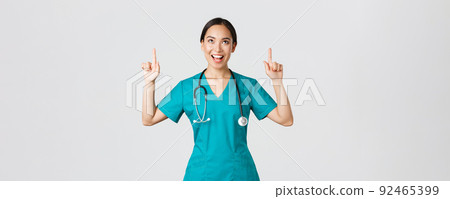 Covid-19, healthcare workers, pandemic concept. Amused cheerful asian female nurse, doctor or surgeon in scrubs pointing fingers and looking up with satisfied face, standing white background 92465399