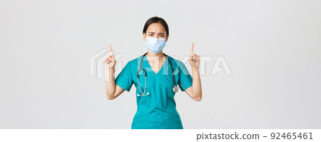 Covid-19, coronavirus disease, healthcare workers concept. Gloomy disappointed asian nurse, physician in medical mask and scrubs, pointing fingers up and frowning perplexed, look frustrated 92465461