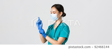 Covid-19, coronavirus disease, healthcare workers concept. Professional smiling asian female nurse, physician in scrubs and medical mask put on rubber gloves for checkup, patient examination 92466144