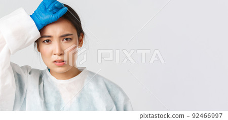 Covid-19, coronavirus disease, healthcare workers concept. Young tired asian female doctor take-off personal protective equipment and wipe sweat, looking exhausted after long shift in hospital 92466997